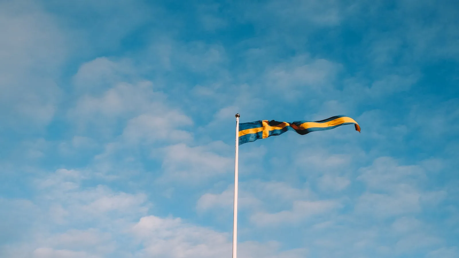 Sweden Visa Website 2025: New Official Portal Launched (March 5th) 1 A Swedish flag waves atop a flagpole under a clear blue sky with scattered clouds.